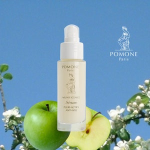 May include: A clear glass bottle of serum with a white pump top. The bottle is labelled "Pomone Paris" and "Munificence Serum Pluri-Actifs Anti-Age". The bottle is sitting on a white surface with a green apple and a half apple in the foreground.
