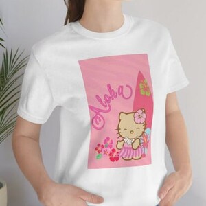 May include: White t-shirt with a pink graphic featuring Hello Kitty wearing a lei and holding a surfboard. The graphic also includes the text "Aloha" and pink flowers.
