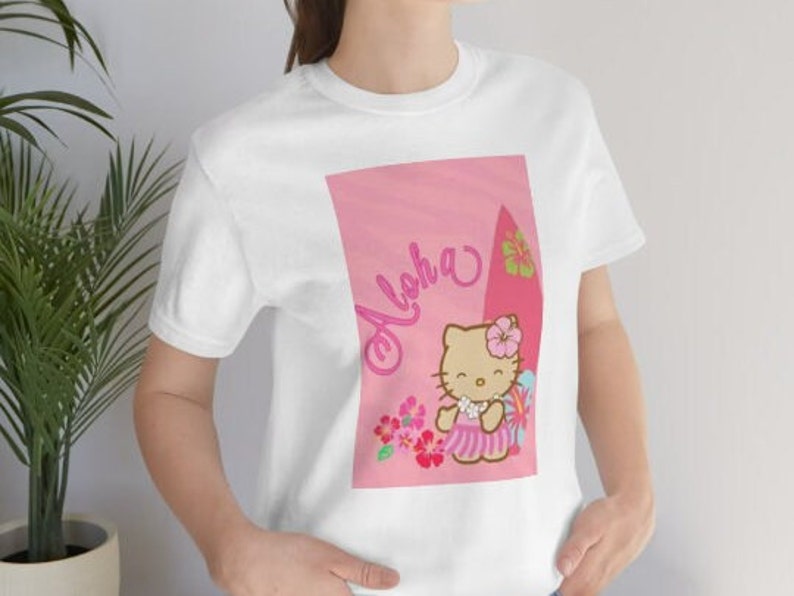 May include: White t-shirt with a pink graphic featuring Hello Kitty wearing a lei and holding a surfboard. The graphic also includes the text "Aloha" and pink flowers.