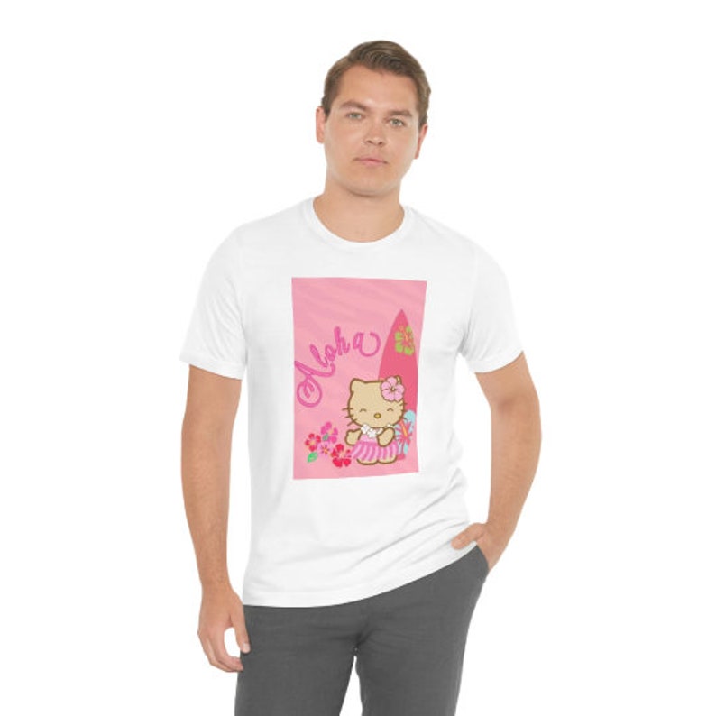 May include: White t-shirt featuring a pink graphic of Hello Kitty on a surfboard with the word "Aloha" in pink lettering.