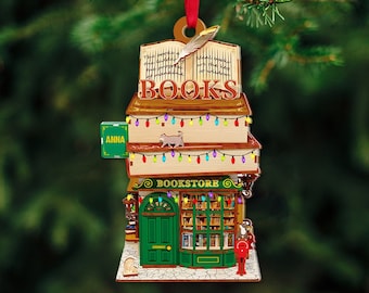 May include: A wooden Christmas ornament shaped like a bookstore with a sign that says "BOOKSTORE" and a sign that says "ANNA". The ornament has a stack of books on top with a sign that says "BOOKS" and a feather on top of the stack. The ornament has string lights and a cat on the top book.