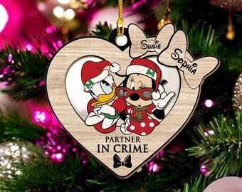 Puede incluir: Un adorno navideño de madera con forma de corazón con una imagen de dos personajes de dibujos animados, un pato y un ratón, que llevan gorros de Papá Noel y gafas de sol. El adorno tiene el texto "Socios en el crimen" y los nombres "Susie" y "Sophia" escritos en él.