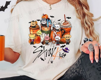 May include: Cream-colored t-shirt with a Halloween-themed graphic featuring cartoon characters, pumpkins, and the text "Stray Kids." An orange mug with a skeleton design is held in the hand. The overall theme is Halloween and fandom.