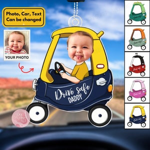 May include: A blue car shaped car air freshener with a yellow roof and a baby's face in the driver's seat. The car has the text "Drive Safe Daddy" on the side. The air freshener is hanging from a rearview mirror. There are several other car air fresheners in different colors in the background.