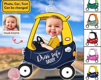 May include: A blue car shaped car air freshener with a yellow roof and a baby's face in the driver's seat. The car has the text "Drive Safe Daddy" on the side. The air freshener is hanging from a rearview mirror. There are several other car air fresheners in different colors in the background.