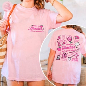May include: A light pink t-shirt with the text "Come on Teacher, Let's go to School!" in pink. The back of the shirt features various school-themed graphics and text, including an apple, globe, and pencil.