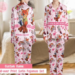 K&ouml;nnte beinhalten: Rosa Satin-Pyjama-Set mit einem sich wiederholenden Muster aus Zeichentrickfiguren, Herzen und dem Text "MITCHELL EMILY". Das lang&auml;rmlige Oberteil und die Hose enthalten eine gro&szlig;e Grafik eines Fuchses und eines Hasen. Der Text "Custom Name" und "All-over Print Satin Pajamas Set" sind ebenfalls sichtbar.