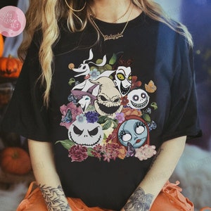 May include: Black t-shirt featuring a colorful graphic of characters from "The Nightmare Before Christmas" surrounded by flowers and butterflies. The design includes Jack Skellington, Sally, and other iconic characters.