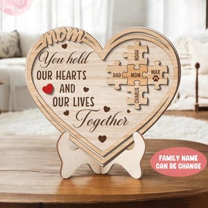 May include: Wooden heart-shaped plaque with a message that reads "You hold our hearts and our lives together". The plaque has a puzzle piece design with the words "Dad", "Mom", "Noah", "Charlie", and "Max" on the pieces. The plaque is on a stand and the text "Family Name Can Be Change" is at the bottom.