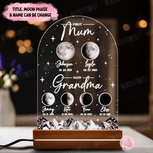 May include: A white acrylic night light with a moon phase design. The light features a title that says "First Mum - Now Grandma" and includes the names of family members with their birth dates. The light is illuminated and sits on a wooden base.