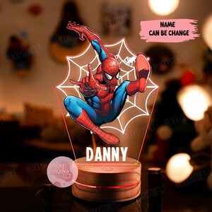 Op de afbeelding: Een 3D-geprint nachtlampje met een rood en blauw Spiderman-personage met een webontwerp. Het nachtlampje heeft een houten basis en de naam "DANNY" is op de voorkant gedrukt. De tekst "NAME CAN BE CHANGE" is in roze op een witte achtergrond geschreven.