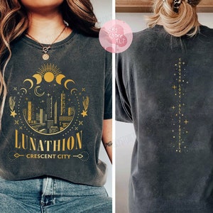 May include: Black t-shirt with a gold graphic of a crescent moon, stars, and a cityscape with the text "Lunathion Crescent City". The back of the shirt has gold text that reads "Through Love, All Is Possible".