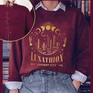 May include: Burgundy sweatshirt with a gold graphic design of a crescent moon, stars, and a city skyline. The text "Lunathion Crescent City" is printed below the graphic.