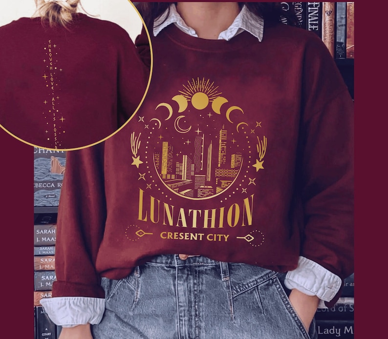 May include: Burgundy sweatshirt with a gold graphic design of a crescent moon, stars, and a city skyline. The text "Lunathion Crescent City" is printed below the graphic.