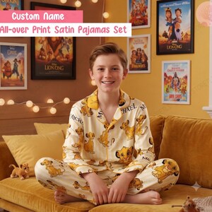 May include: A long-sleeved button-down shirt and matching pants set made of cream-colored satin fabric. The pajamas feature an all-over print of the character Simba from The Lion King, interspersed with the custom name Michael in black text.