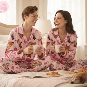 K&ouml;nnte beinhalten: Passende rosa Pyjama-Sets mit einem Cartoon-Fuchs- und Herzmuster. Die Pyjamas umfassen ein Hemd mit Kn&ouml;pfen und eine Hose. Das Paar sitzt auf einem Bett und h&auml;lt wei&szlig;e Tassen.