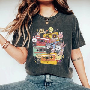 Puede incluir: Una camiseta gris oscuro con un estampado gráfico de animales de dibujos animados y cintas de casete. El gráfico presenta el texto "Circus", "No Easy", "Manliest", "Miroh" y "A Side".