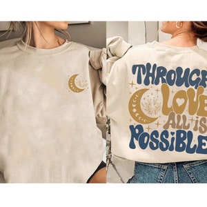 May include: A beige sweatshirt with a crescent moon and stars design on the front and the text "Through Love All is Possible" in blue and gold lettering on the back.