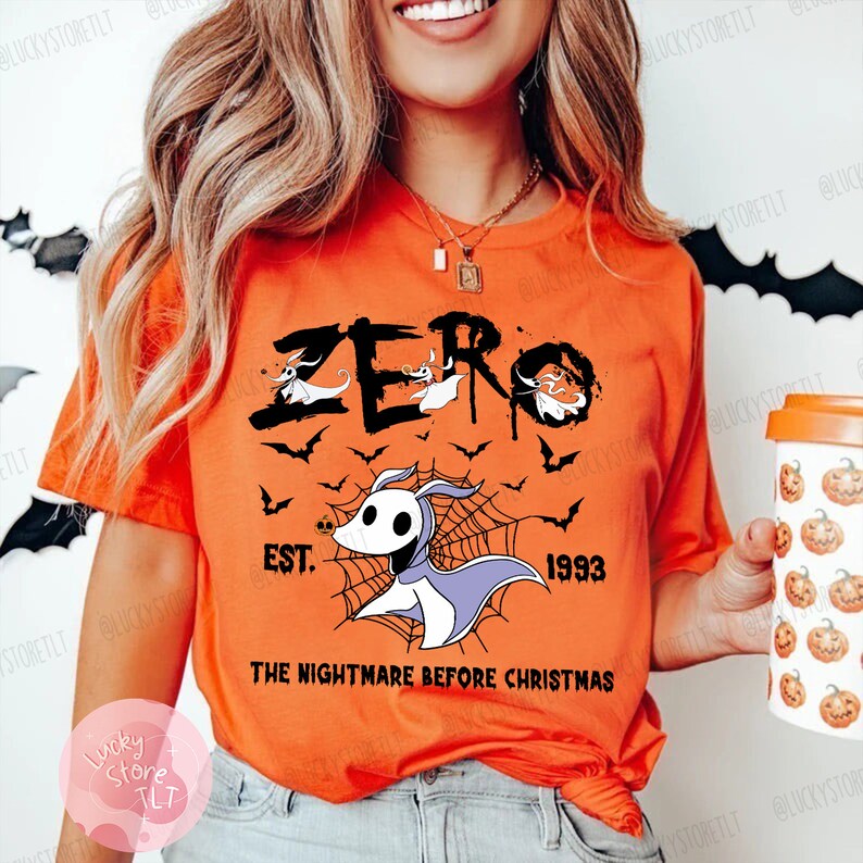 May include: Orange t-shirt featuring the word "ZERO" with a graphic of Zero from "The Nightmare Before Christmas." The design includes bats, a spiderweb, and the text "EST. 1993." The shirt is a short-sleeved crew neck.