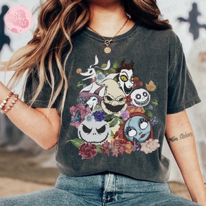 May include: Dark gray t-shirt featuring a colorful graphic of characters from The Nightmare Before Christmas, surrounded by flowers and butterflies. The design includes Jack Skellington, Sally, and other iconic figures. The shirt is a comfortable, casual style.