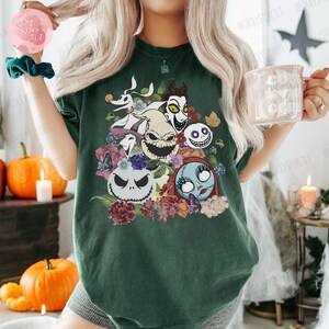 May include: Dark green t-shirt featuring a colorful graphic of characters from The Nightmare Before Christmas, including Jack Skellington, Sally, and Zero, surrounded by flowers and butterflies. The shirt is a comfortable, casual style.