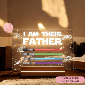 May include: A clear acrylic sign with a white background and the text "I AM THEIR FATHER" in white. The sign has a starry night sky background with four lightsabers and spaceships. The lightsabers are labeled with the names "JULA", "JANET", "TIANA", and "JOSHUA". The sign is on a wooden base. The text "NAME & SABER CAN BE CHANGE" is in pink at the bottom of the image.