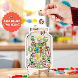 Pode incluir: Um expositor de madeira em forma de jarro com um suporte, cheio de fichas de personagens coloridas. O jarro está rotulado como "AIDYN'S SUPER JAR" e inclui um autocolante "Best Seller". Uma mão está a colocar uma ficha no jarro. Tamanho L. Suporte incluído.