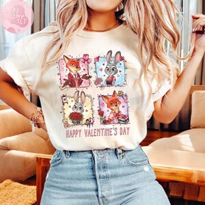 Pu&ograve; includere: T-shirt color crema con una grafica colorata di personaggi dei cartoni animati a tema San Valentino. Il design include una volpe e un coniglio in varie pose, circondati da cuori e fiori. Il testo "HAPPY VALENTINE'S DAY" &egrave; stampato sotto.