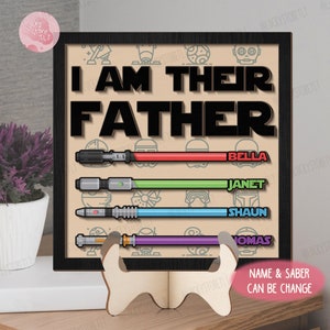 Pode incluir: Um sinal quadrado com moldura preta e o texto "I AM THEIR FATHER" em letras brancas grandes.  Abaixo do texto, h&aacute; quatro imagens de sabres de luz, cada uma com uma cor diferente e um nome abaixo: Bella, Janet, Shaun e Thomas.  O sinal est&aacute; em um suporte de madeira.