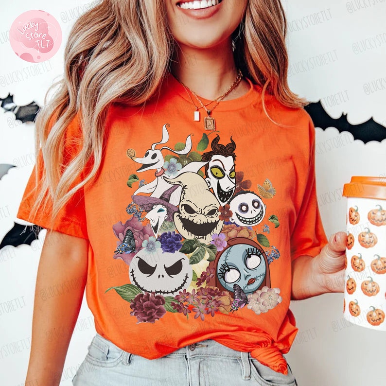 May include: Orange t-shirt featuring a colorful graphic of characters from The Nightmare Before Christmas, surrounded by flowers and butterflies. The design includes Jack Skellington, Sally, and other iconic figures. The shirt is a short-sleeved crew neck.