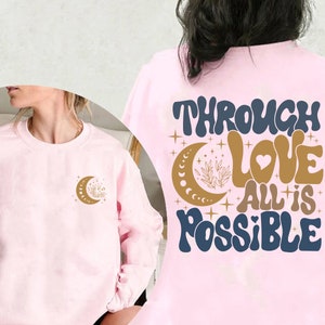 May include: Pink sweatshirt with the text "Through Love All is Possible" in blue and gold lettering with a crescent moon and stars. The front of the sweatshirt has a smaller crescent moon with stars in gold.