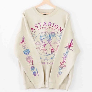 Puede incluir: Una sudadera beige con un estampado gráfico de un hombre con el pelo largo y una espada. El gráfico está en tonos de rosa y azul e incluye el texto "Astarion Approves" y "Your's Game".
