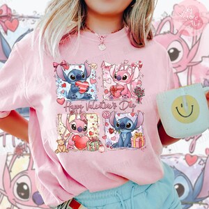 Puede incluir: Camiseta rosa claro con un gr&aacute;fico de cuatro paneles de personajes de dibujos animados sosteniendo corazones y regalos. El texto "Happy Valentine's Day" est&aacute; impreso debajo de los dos paneles superiores. En la imagen se sostiene una taza blanca con una carita sonriente amarilla.