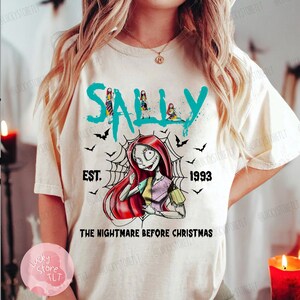 May include: Cream-colored t-shirt featuring a graphic of Sally from "The Nightmare Before Christmas." The design includes the name "SALLY" in teal, the year "1993," and the phrase "THE NIGHTMARE BEFORE CHRISTMAS." Black bats and a spiderweb complete the design.