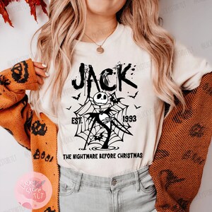 May include: Cream-colored t-shirt featuring a black graphic of Jack Skellington from "The Nightmare Before Christmas." The design includes the word "JACK," bats, a spiderweb, and the text "EST. 1993." The shirt is part of a Halloween-themed collection.