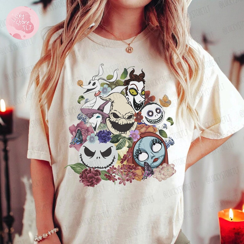 May include: Cream-colored t-shirt featuring a colorful graphic of characters from "The Nightmare Before Christmas" surrounded by flowers and butterflies. The design includes Jack Skellington, Sally, Zero, and other iconic characters. A unique and whimsical design.