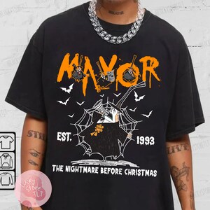 May include: Black t-shirt featuring a graphic of the Mayor from "The Nightmare Before Christmas." The design includes the word "MAYOR" in orange, bats, and the text "EST. 1993." The shirt is a classic crew neck.