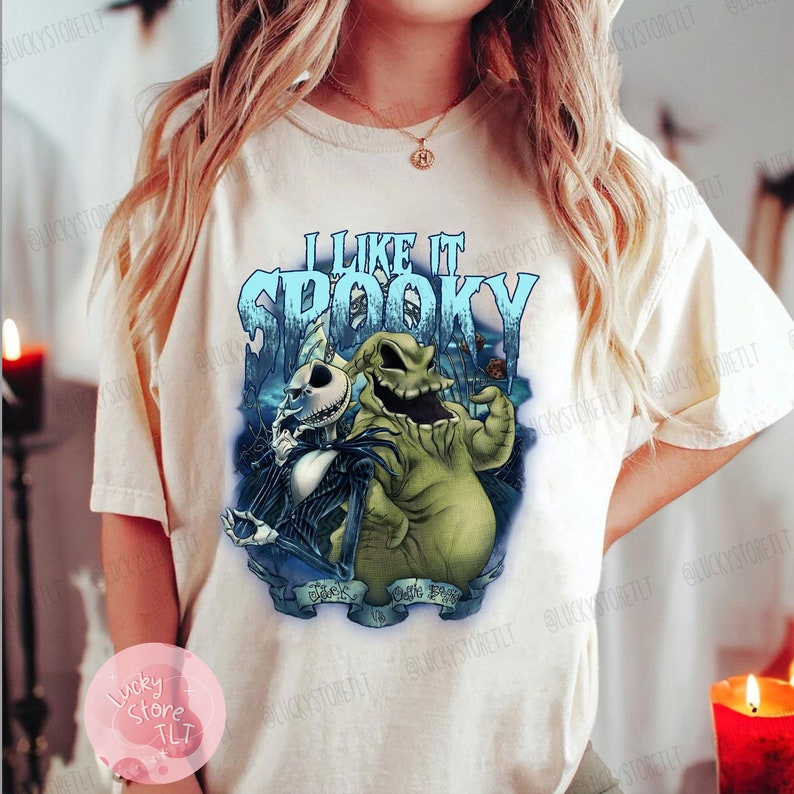 May include: Cream-colored t-shirt featuring a graphic of Jack Skellington and Oogie Boogie from The Nightmare Before Christmas. The text "I Like It Spooky" is in blue, with a spooky background. The shirt is a casual wear item.