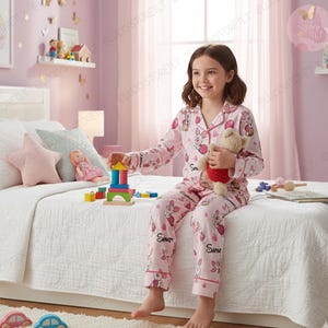 May include: A young person wearing pink pajamas with a Winnie the Pooh pattern, holding a teddy bear. The pajamas have a button-down top and matching pants. Colorful wooden blocks are on the bed, along with a star-shaped pillow.