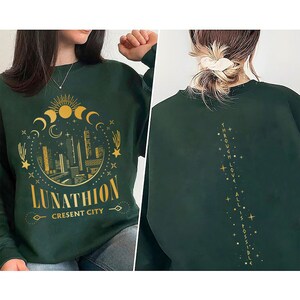 May include: A dark green sweatshirt with a gold graphic of a crescent moon, stars, and a cityscape with the text "Lunathion Crescent City". The back of the sweatshirt has the text "Through Love All Is Possible" in gold.