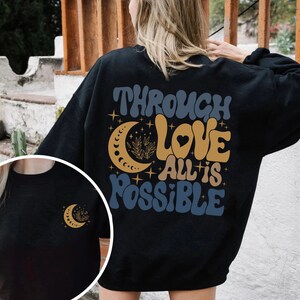 May include: Black sweatshirt with a graphic design featuring a crescent moon, stars, and the text "Through Love All is Possible".