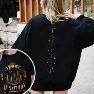 May include: Black sweatshirt with a gold graphic on the back that says "Through love, all is possible." The front of the sweatshirt has a gold graphic of a crescent moon, stars, and a cityscape with the text "Lunathion Crescent City".