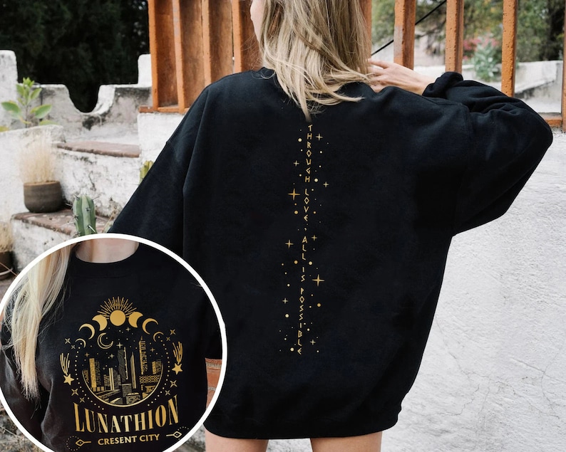 May include: Black sweatshirt with a gold graphic on the back that says "Through love, all is possible." The front of the sweatshirt has a gold graphic of a crescent moon, stars, and a cityscape with the text "Lunathion Crescent City".