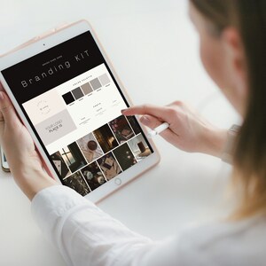 May include: A person is using a tablet displaying a "Branding Kit" layout. The screen shows a logo design, color palette, and sample images. The tablet is held in a light-colored case, and a stylus is used to interact with the screen.