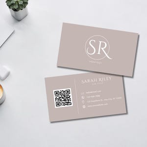 May include: A light brown business card with a white circle containing the letters "SR" and the text "SARAH RILEY" below. The card also includes a QR code, contact information, and the text "SARAH RILEY Skin Care".