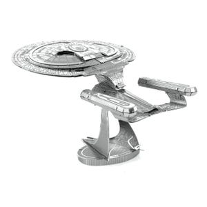 Puede incluir: Maqueta metálica plateada de una nave espacial, con un cuerpo principal circular y dos estructuras en forma de ala. El modelo es intrincadamente detallado y se apoya en un pedestal, mostrando un diseño futurista. El modelo mide unos 30 cm de largo.