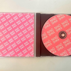 May include: A CD case and disc with a pink and white repeating pattern of the words "NINU SHKA". The CD is in a black case. The disc is pink with the same text.