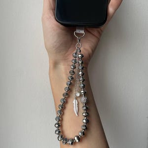 Peut inclure: Un porte-téléphone à perles grises, avec des perles à facettes et des breloques décoratives. Le porte-téléphone comprend une plume argentée et une petite perle scintillante. Le porte-téléphone est attaché à un smartphone noir.