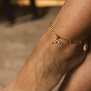 May include: A gold-toned anklet featuring a delicate chain with small evenly spaced beads and a central charm shaped like a whale tail, which is encrusted with tiny clear stones. A second, simpler gold-toned chain is visible around the other ankle.