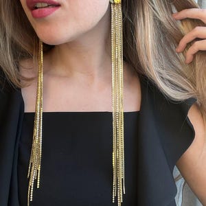 May include: A pair of long, gold-toned fringe earrings featuring a cluster of yellow and clear crystals at the top. Multiple thin, shimmering metallic chains hang down from the crystal base, creating a cascading effect that reaches the chest.